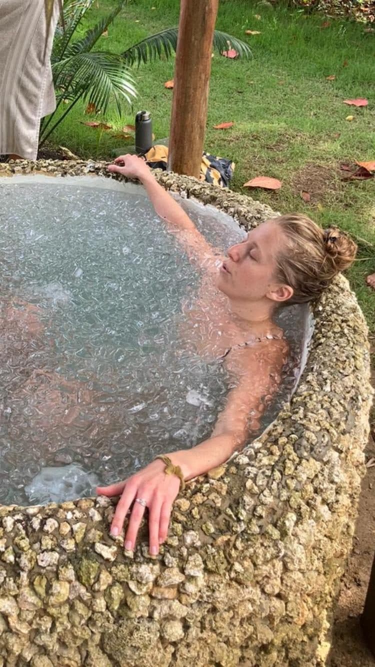 A person resting in an outdoor ice bath, in a round stone basin surrounded by grass.