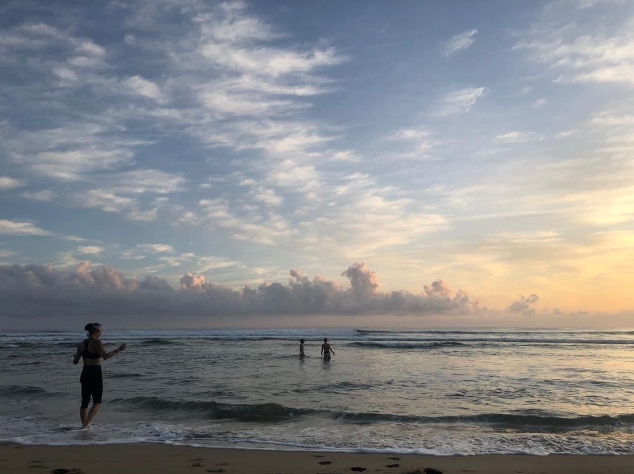 People wading into the sea at dawn beneath a pale, cloud-streaked sky.